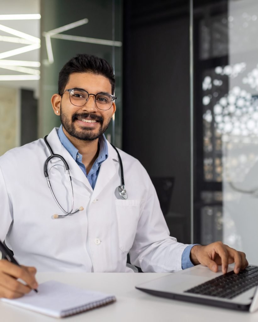 Smiling male healthcare professional in white lab coat using computer at his desk in a contemporary clinic setting.